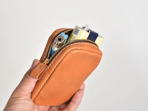 職人手作皮革菸盒收納包，顏色為溫暖的橘棕色，材質為真皮。包體縫線明顯，配有金色拉鍊及同色系的皮革拉頭。包內裝有香菸盒及打火機。