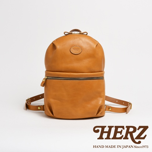 職人手作皮革大開口圓角後背包 R-18，採用溫暖的焦糖色皮革，搭配銀色拉鍊和金屬扣環，設計簡約，質感細緻，適合日常搭配。