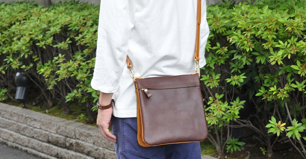 職人手作皮革翻轉雙色直式斜背包，主體為深棕色皮革，側邊為淺棕色皮革，配有拉鍊和可調節的皮革斜背帶。