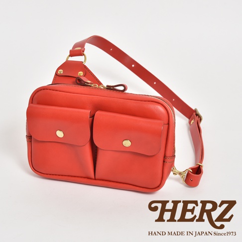 紅色皮革雙口袋斜背包，具有兩個前方翻蓋口袋，附有黃銅釦。背包採用拉鍊開合，配有可調節的皮革肩帶。