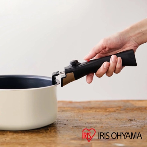 一只手正將一個黑色可拆卸鍋柄，裝到一個淺奶油色鍋子的邊緣。鍋柄上有一個黑色旋鈕和一個紅色按鈕，接口處有金色金屬部件。