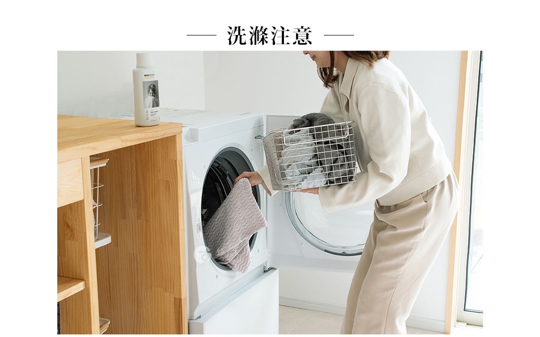 一位女子正將一條米色波浪紋毛巾放入打開的洗衣機滾筒內，手中還拿著一個裝有灰黑色毛巾的金屬洗衣籃。