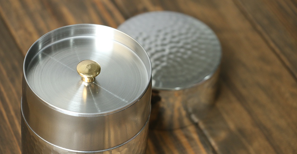 不鏽鋼錘目紋茶葉罐，帶有金色圓形提把，罐身和罐蓋皆為銀色金屬材質，罐身下方有明顯的錘擊紋路。