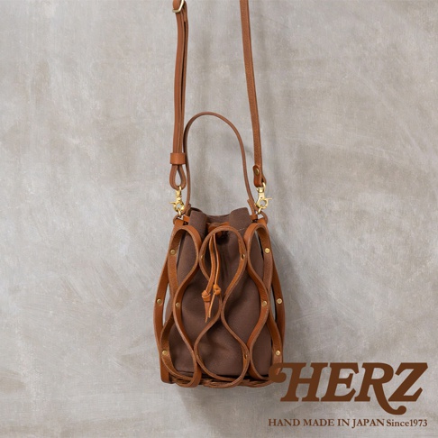 日本 HERZ 職人手作皮革 Organ 編織水桶包 G-98，棕色帆布包體搭配編織皮革外罩，附有長短提把和可拆卸斜揹帶，HERZ LOGO 印製於右下角。