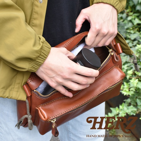 職人手作的棕色皮革腰掛斜背包，包款簡約，有拉鍊主袋與前置拉鍊口袋，搭配著橄欖綠外套與淺色褲子，背景為綠色植被。