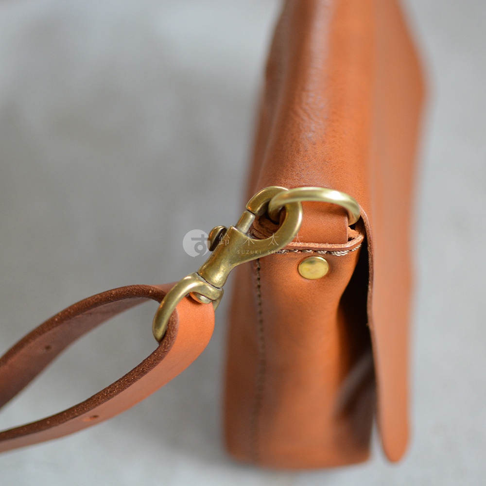 特寫鏡頭展示了職人手作的棕色皮革斜背包，其金屬扣環與皮革背帶連接，細膩的縫線和自然的皮革紋理清晰可見。
