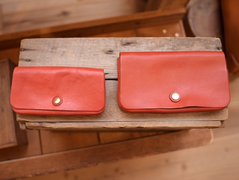 兩款橘紅色皮革長夾，皆配有金色按扣。左邊較小，右邊較大，均置於木製層架上。