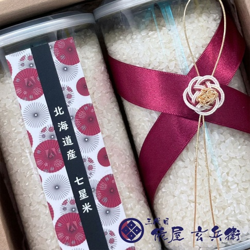 兩個裝著白色米粒的透明圓柱形容器並排放置在禮盒中，左側容器貼有印著紅色圓形圖案與北海道產七星米字樣的包裝紙，右側容器則綁著深紅色緞帶與精緻的金色結飾，右下角標示有三代目俵屋玄兵衛的品牌商標。