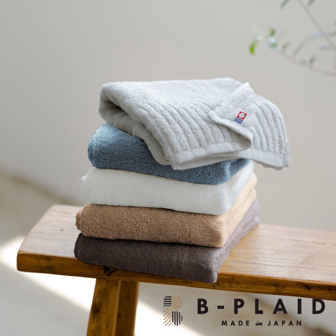 一疊五條今治毛巾，顏色由上至下為淺灰、藍色、白色、褐色、深棕色。淺灰色毛巾帶有明顯的直條紋理，並有一個紅色、藍色和白色標誌。