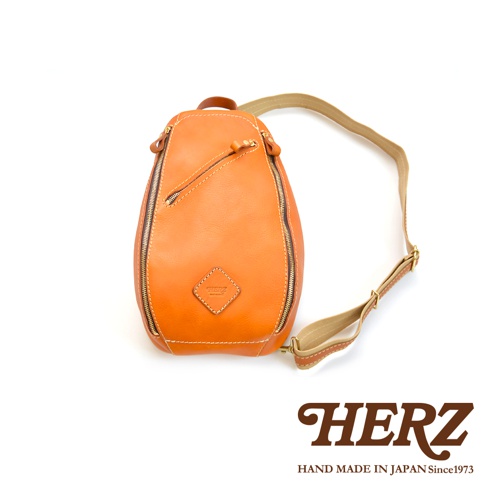 一皮革製斜背包，呈水滴狀，表面光滑，帶有拉鍊開合，左側有一傾斜拉鍊口袋，前方中央有一個菱形皮革標籤，附有可調節的帆布與皮革混搭肩帶，顏色為橘棕色。