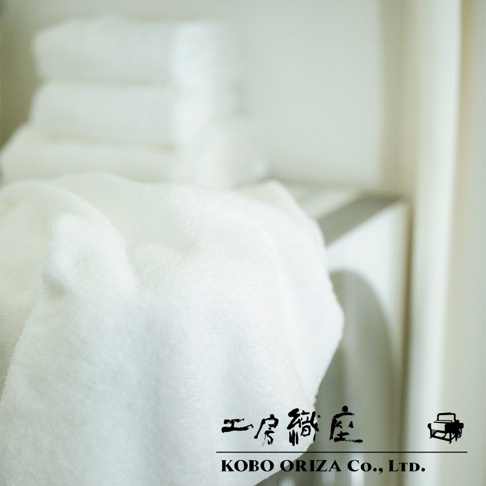 一條柔軟的白色匹馬棉毛巾，質地蓬鬆，疊放在背景堆疊的白色毛巾之上。