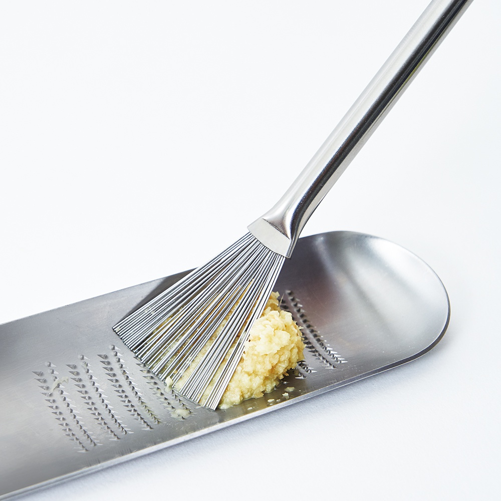 日製料理小刷，由不鏽鋼製成，設有細長的金屬刷毛，用於刮取蒜泥。小刷架置於一個橢圓形的銀色不鏽鋼研磨器上，研磨器表面有細小的齒紋，中間有一小堆黃色蒜泥。