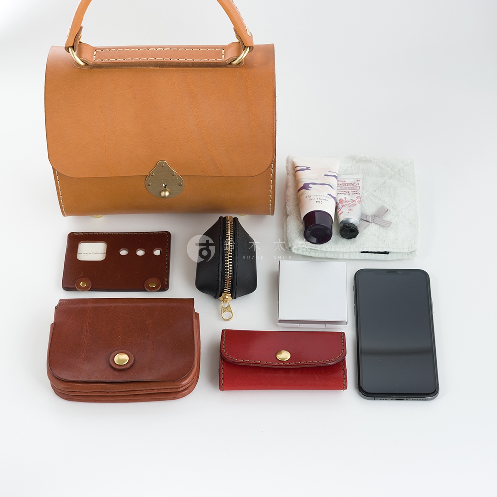 職人手作皮革復古杜勒斯手提包，為淺棕色，配有金色金屬鎖扣和手柄。包旁散落著一些物品，包括一個棕色皮革卡夾、一個黑色拉鍊零錢包、一個紅色皮革零錢包、一個手機、兩管護膚品和一塊淺綠色毛巾。