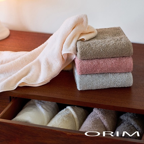一張深色木製家具上擺放著多條 ORIM 品牌的柔軟毛巾，上方堆疊著棕色、粉色與灰色的厚實毛巾，側面垂掛著一條淺米色毛巾，下方的抽屜則整齊收納著多條不同色系的捲裝毛巾，展現出毛巾澎厚柔軟的細緻觸感。