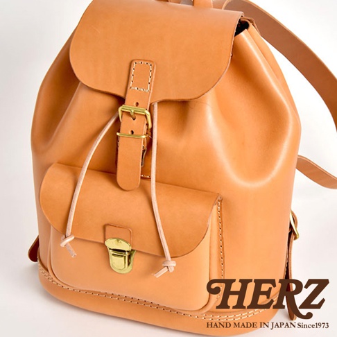 職人手作皮革經典後背包，天然皮革材質，原色調，設有翻蓋、束繩口袋、金屬扣環及可調式肩帶。