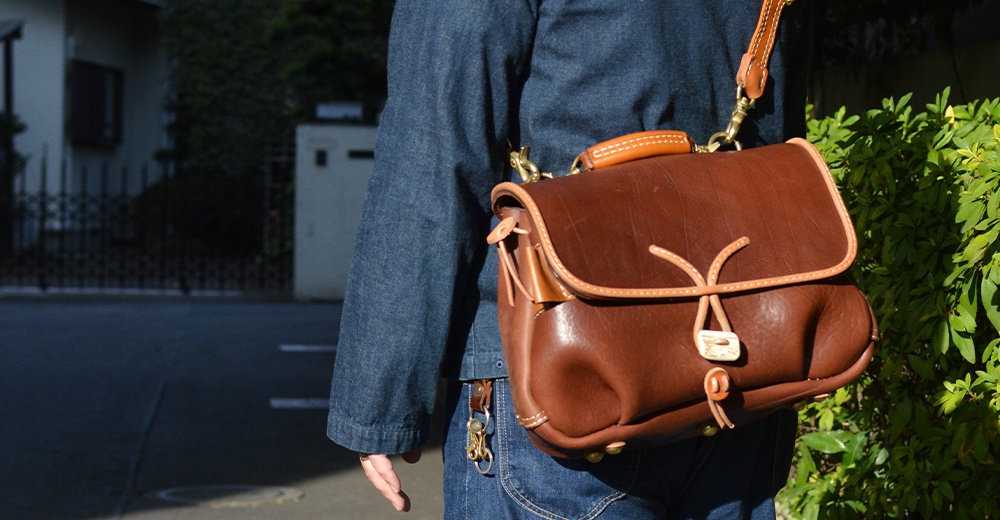 棕色皮革鹿角扣繩郵差包，具備皮革提把與可調式肩帶，包身有抽繩設計，搭配金屬扣環，適合日常攜帶。