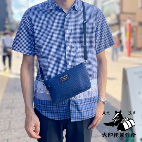 深藍色刺子織隨身斜背包，斜挎於身穿藍色格紋襯衫的男子身上，包包上有品牌標籤。
