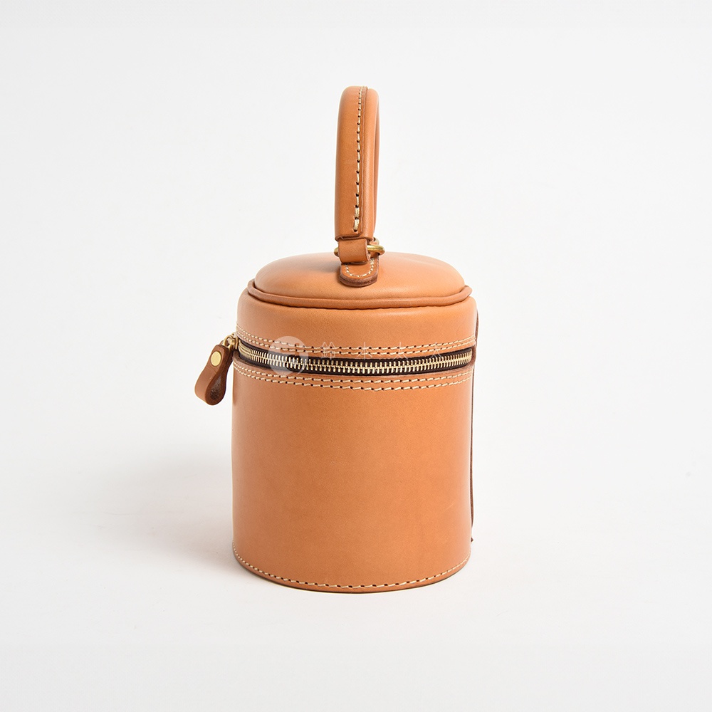 一個圓筒狀的淺棕色皮革化妝手提包，頂部有提把，側面有拉鍊開合，手工縫線清晰可見。