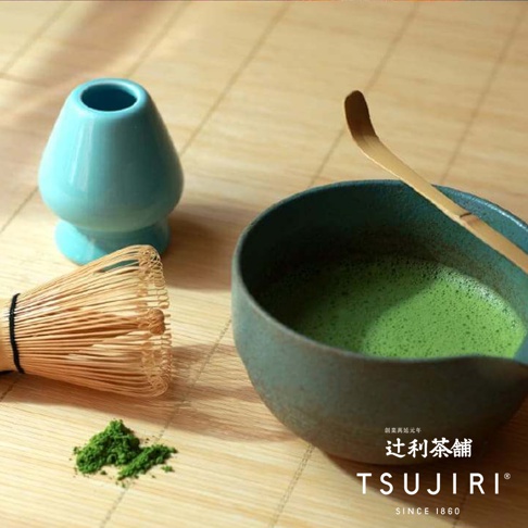 這張照片展示了傳統抹茶的沖泡器具，包含一個擺放在淺藍色陶瓷刷架旁的竹製茶筅、一小堆鮮綠色的抹茶粉，以及一碗深色粗陶茶碗，碗內裝有已打散泡沫的綠色抹茶，旁邊斜放著一支木製茶杓，右下方印有辻利茶舖的品牌名稱與標誌。