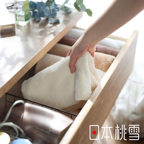 一隻手正從木質抽屜中拿取一條淺米色的日本桃雪精梳棉浴巾，抽屜內整齊收納著多條白色與棕色的柔軟浴巾，左下角可見一台吹風機，整體畫面呈現溫馨且居家收納的舒適氛圍。