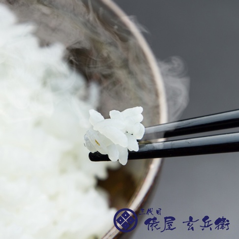 一雙黑色的筷子夾著一團熱騰騰的白米飯，米飯飽滿晶瑩，散發出陣陣白煙。背景是裝滿米飯的碗，碗邊緣有深色裝飾，整體呈現溫暖樸實的氛圍。