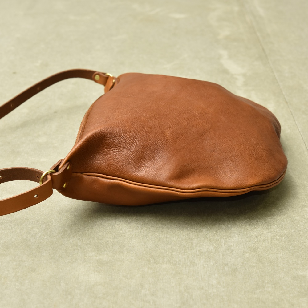 棕色皮革製成的單肩半月包，具有可調節的皮革背帶和金色金屬配件。包身呈現自然的皮革紋理，線條簡潔圓潤。
