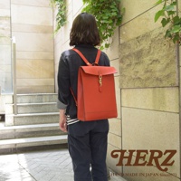 身穿黑色外衣的模特背著一個橘紅色皮革後背包，背包設計簡約，具有紙袋的輪廓，配有金色鉤扣和皮質肩帶，展示了精湛的手工藝。