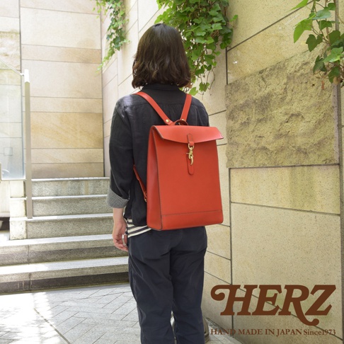 身穿黑色外衣的模特背著一個橘紅色皮革後背包，背包設計簡約，具有紙袋的輪廓，配有金色鉤扣和皮質肩帶，展示了精湛的手工藝。