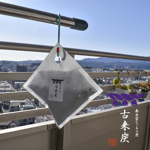 一個方形的白色透氣織物袋，內含黑色炭粒，袋上貼有標籤，標籤上有日文文字及圖案。袋頂有綠色掛繩，掛在金屬欄杆上。背景是城市景觀和遠山。