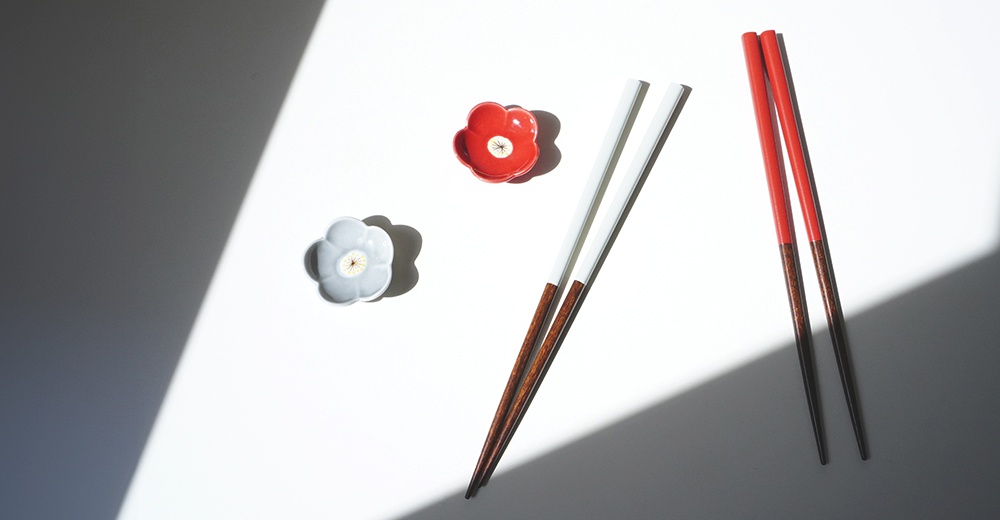 一雙白色筷子與一雙紅色筷子，均為木質，頂端有木紋，另有兩個花朵造型的淺盤，一個為紅色，一個為灰色。