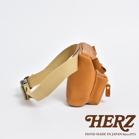 棕色皮革製成的職人手作腰掛斜背包，側邊帶有拉鍊口袋，搭配米色織帶及黑色扣環。