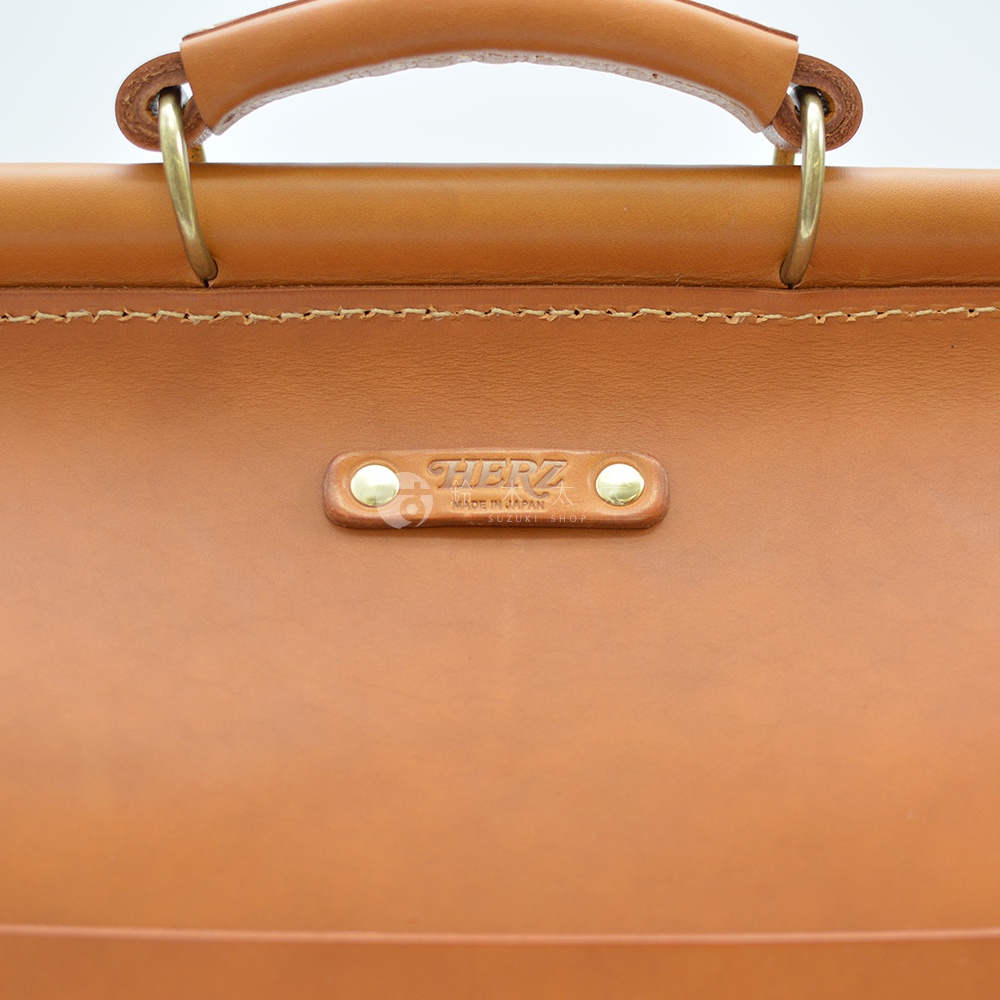 職人手作皮革旅行包 A-102，採用光滑棕色皮革製作，附有圓形提把和金屬扣環。包身中央有壓印品牌標誌的皮革銘牌，標誌下方印有「MADE IN JAPAN」和「SUZUKI SHOP」。