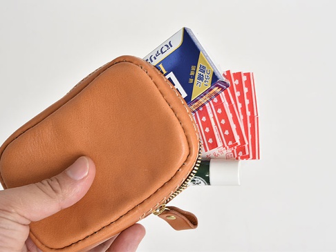 淺棕色皮革菸盒收納包，拉鍊開口，內含一盒藍色包裝的日文菸盒、數張紅色圖案紙片及一支白色護唇膏。