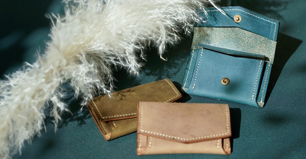 北海道KASANE 職人手作義大利Texas皮革迷你雙折短夾，近距離拍攝，以淺色植物羽毛為前景，搭配深藍色背景，展示了三個不同顏色（藍色、橄欖綠、駝色）的皮革短夾，短夾有精緻的縫線和金屬釦，展現手作文藝風格。