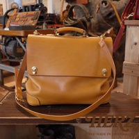 一件棕褐色皮革製作的兩用波士頓包，包身方形，有提把和可拆卸肩帶。包蓋處有兩個金色旋轉鎖扣，邊角處有縫線細節。