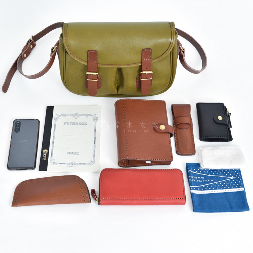 橄欖綠皮革製成的傳統釣魚包，搭配棕色皮革翻蓋和搭扣，與多種皮革配件一起展示，包括棕色筆記本、棕色長夾、紅色長夾、棕色小皮套、黑色錢包、手機和藍色印花手帕。