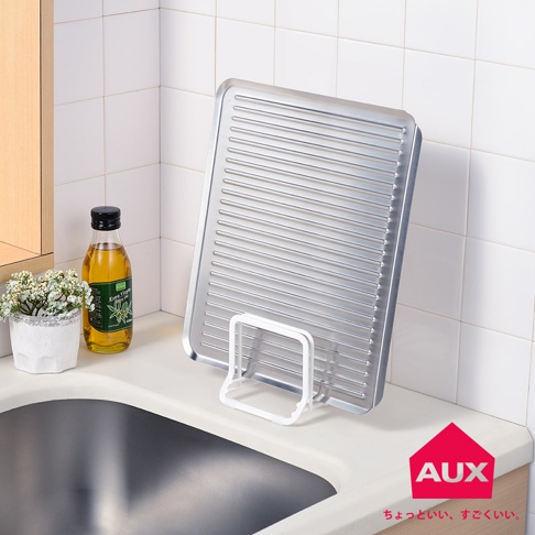 銀色金屬材質的leye 不積水瀝水盤，表面設有多條平行凹槽，以白色支架立於水槽旁，旁邊有一瓶橄欖油和一盆白色小花。