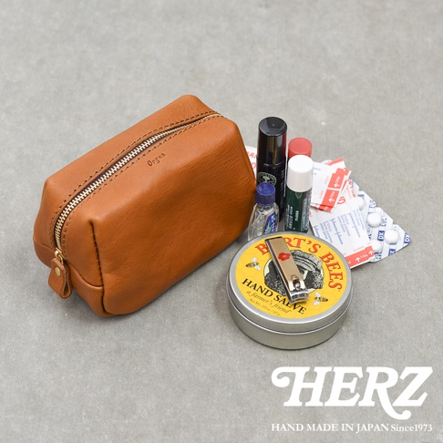 一個棕色皮革拉鍊收納包平放在灰色表面上，包身印有Organ字樣，旁邊擺放著伯特小蜜蜂護手霜圓鐵盒、指甲剪、眼藥水、唇膏及數包藥品和OK繃，右下角印有HERZ品牌標誌及相關文字。