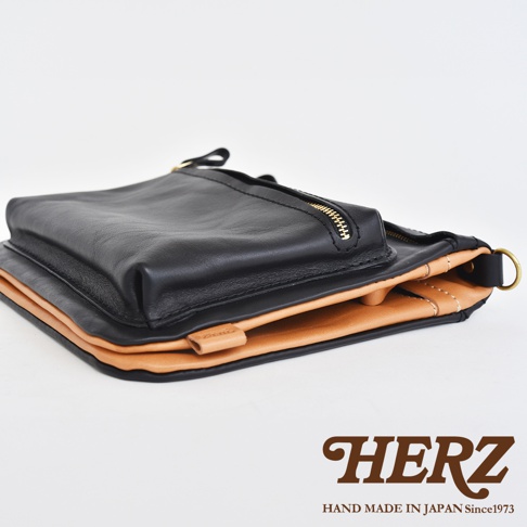 職人手作皮革翻轉雙色直式斜背包，黑色皮革翻蓋搭配原色皮革主體，設有金色拉鍊，展現簡約質感。