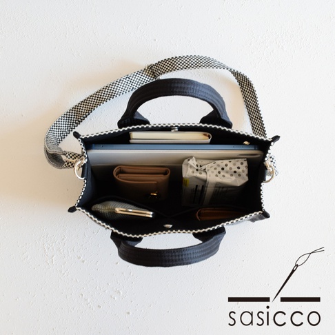 三河木綿刺子織兩用方形托特包，黑色主體搭配白色格紋提帶，包內裝有筆記本、平板電腦、棕色皮夾、手機及其他小物，展現實用收納空間。