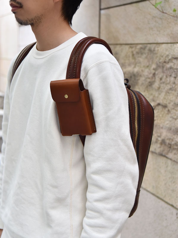 棕色皮革三用手機收納包，附有金屬鈕扣，固定於白色長袖上衣的肩帶上，旁邊為同款式的棕色皮革後背包。