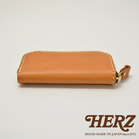 駝色皮革製成的橢圓形拉鍊鑰匙包，有縫線細節和黃銅色拉鍊，底部有 HERZ 標誌和 HAND MADE IN JAPAN Since1973 字樣