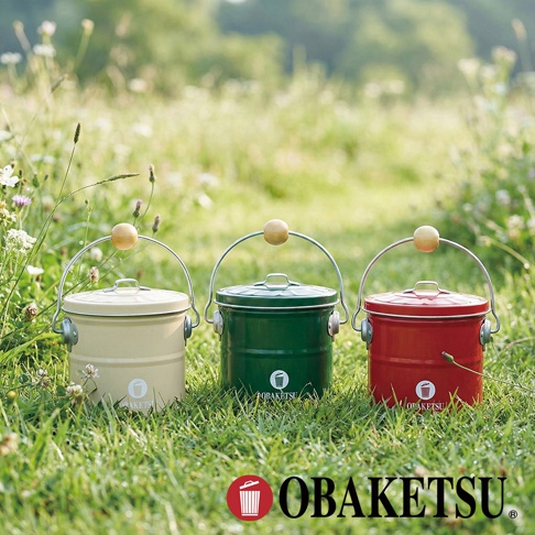 OBAKETSU 品牌的三個手工製蚊香桶，分別為米白色、綠色和紅色，擺放在青翠的草地上，背景是模糊的綠色植物。