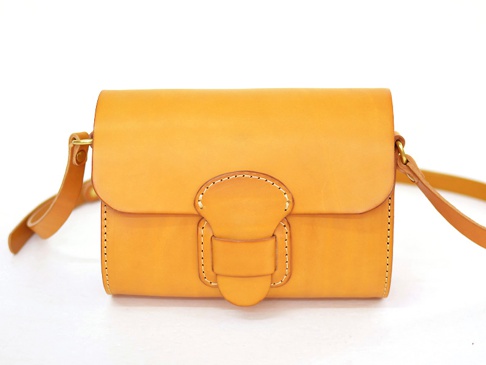 淺棕色皮革馬鞍包，有寬版皮帶和獨特的皮革搭扣設計，車縫線清晰可見，整體呈現簡約風格。