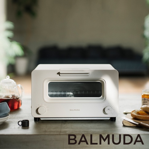 白色 BALMUDA 蒸氣烤麵包機，設有玻璃門和兩個控制旋鈕，擺放在灰色檯面上，背景是模糊的室內環境。