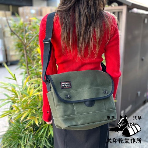 綠色純棉帆布雙扣郵差包，配有黑色織帶和黑色滾邊，包蓋上有品牌標籤。