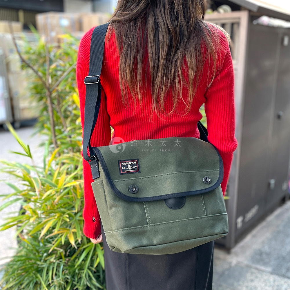 身著紅色毛衣的女性，斜挎著一個墨綠色純棉帆布雙扣郵差包。包包有兩個黑色圓形按扣，一個黑色皮革裝飾，以及一個方形的品牌標籤。