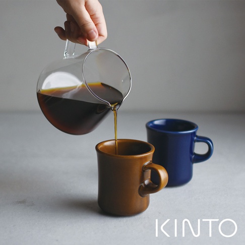 一只手拿著透明玻璃咖啡下壺，正將深棕色咖啡倒入一個淺棕色陶瓷馬克杯中。背景有一藍色陶瓷馬克杯。