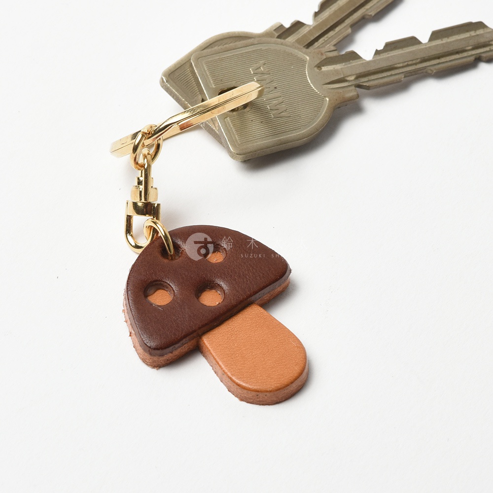 職人手作皮革香菇造型鑰匙圈，深棕色傘蓋有兩處圓孔，搭配淺棕色蕈柄，以金色金屬環和掛鉤連接。