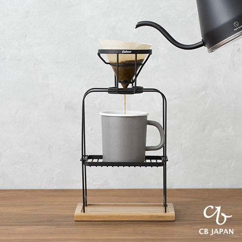 黑色金屬兩段式手沖咖啡架，上層支撐著印有「Qahwa」字樣的濾杯，濾杯中裝有咖啡粉，下方為灰色馬克杯，整體置於木質底座上。